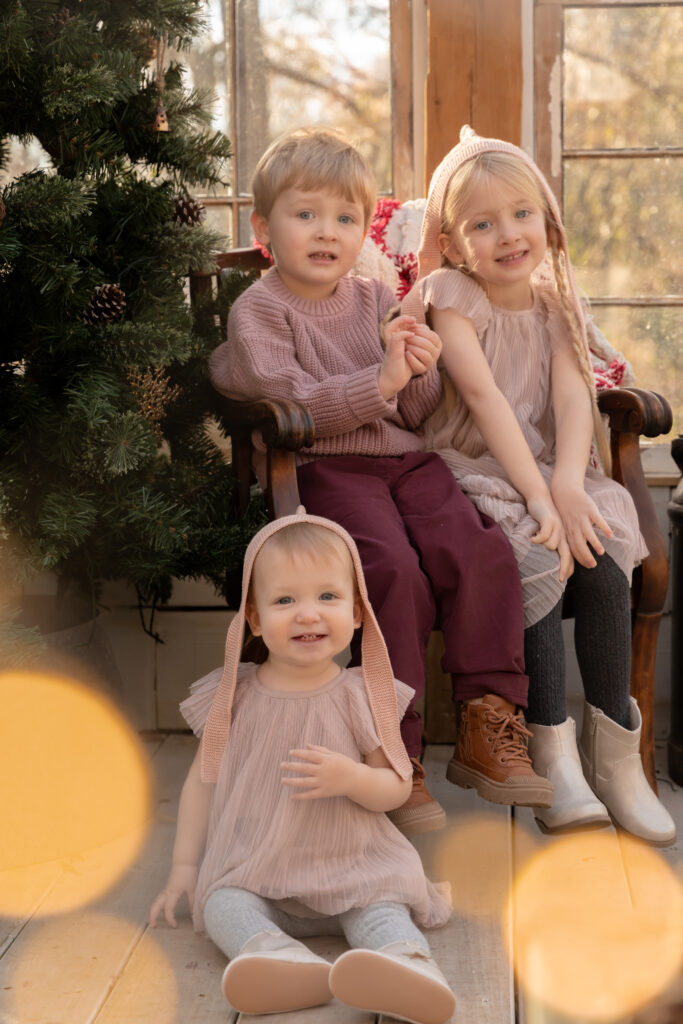 Three children sit together smiling at the camera in a greenhouse decorated for Christmas