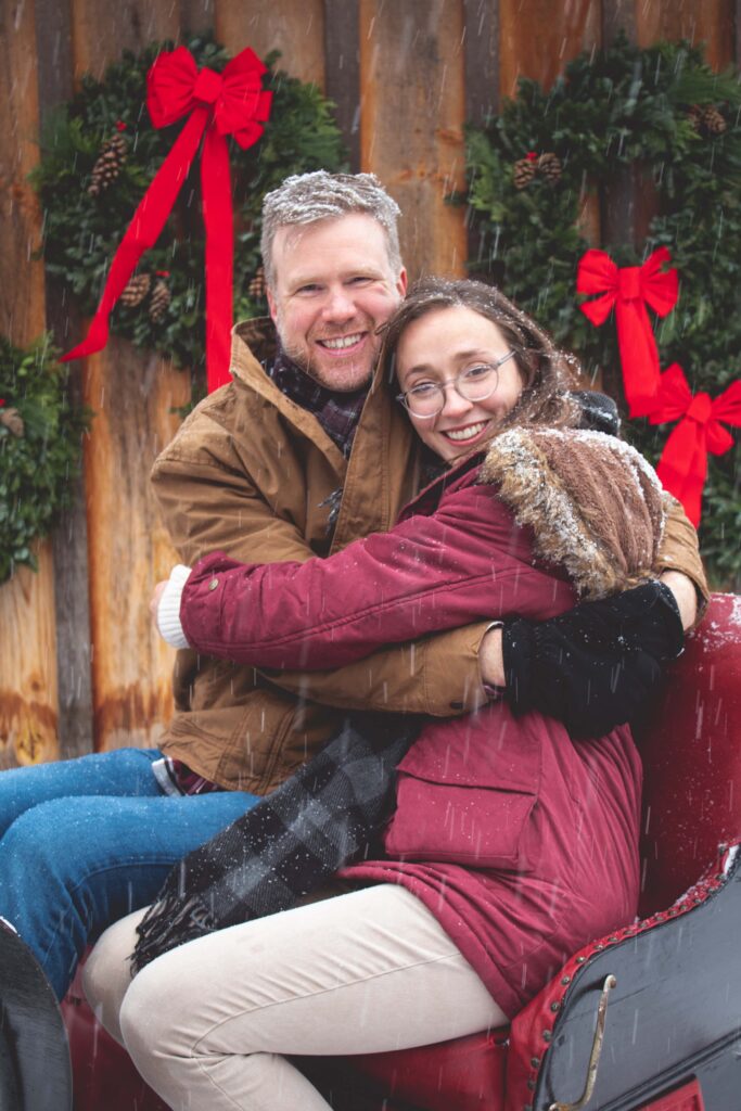 Couple cuddles and smiles at the camera on a sleigh that sits in front of a barn with Christmas wreaths during a snow flurry