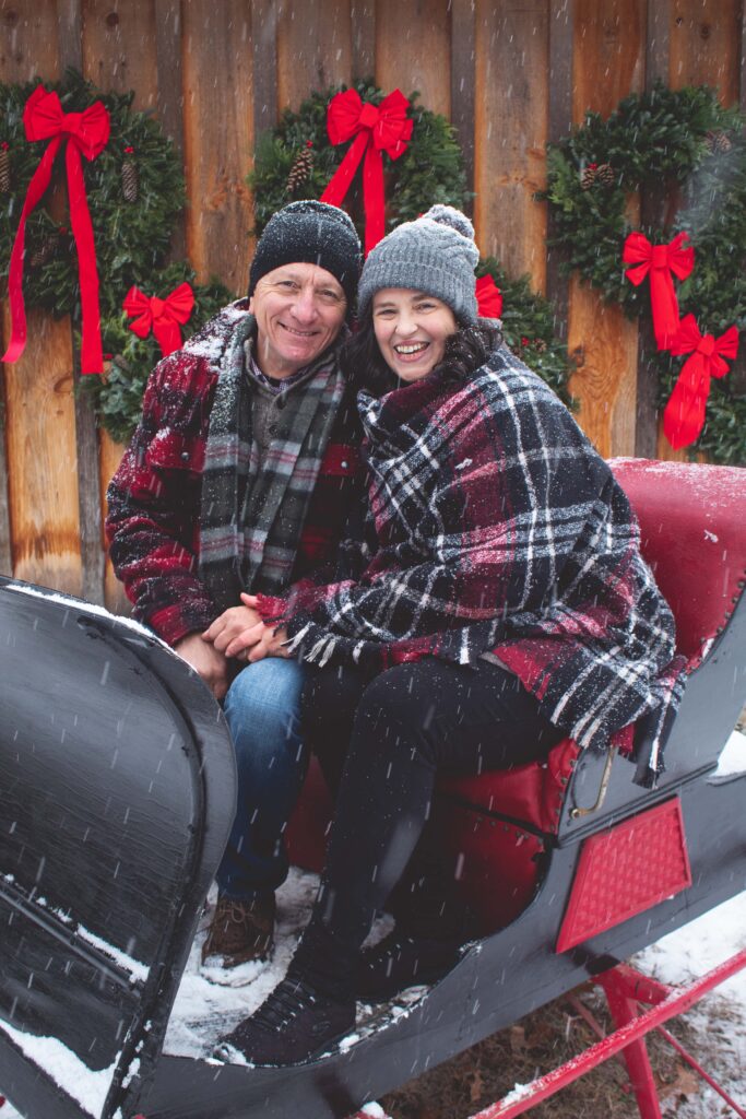 Couple cuddles and smiles at the camera on a sleigh that sits in front of a barn with Christmas wreaths during a snow flurry