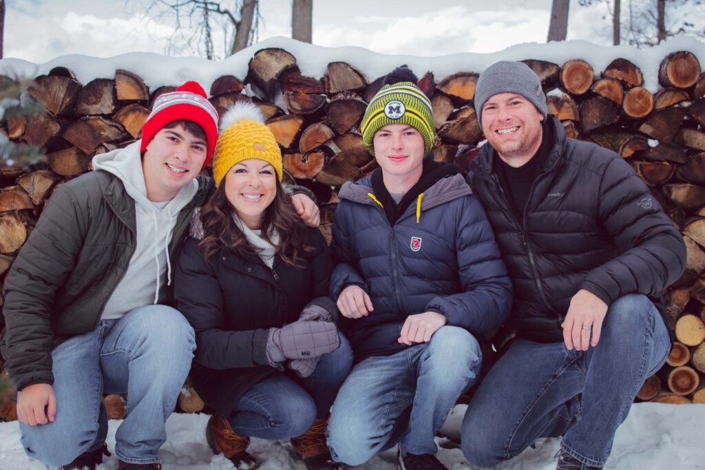 Family of 4 with two teenage boys squats in front of a wood pile in the snow, all wearing winter gear 