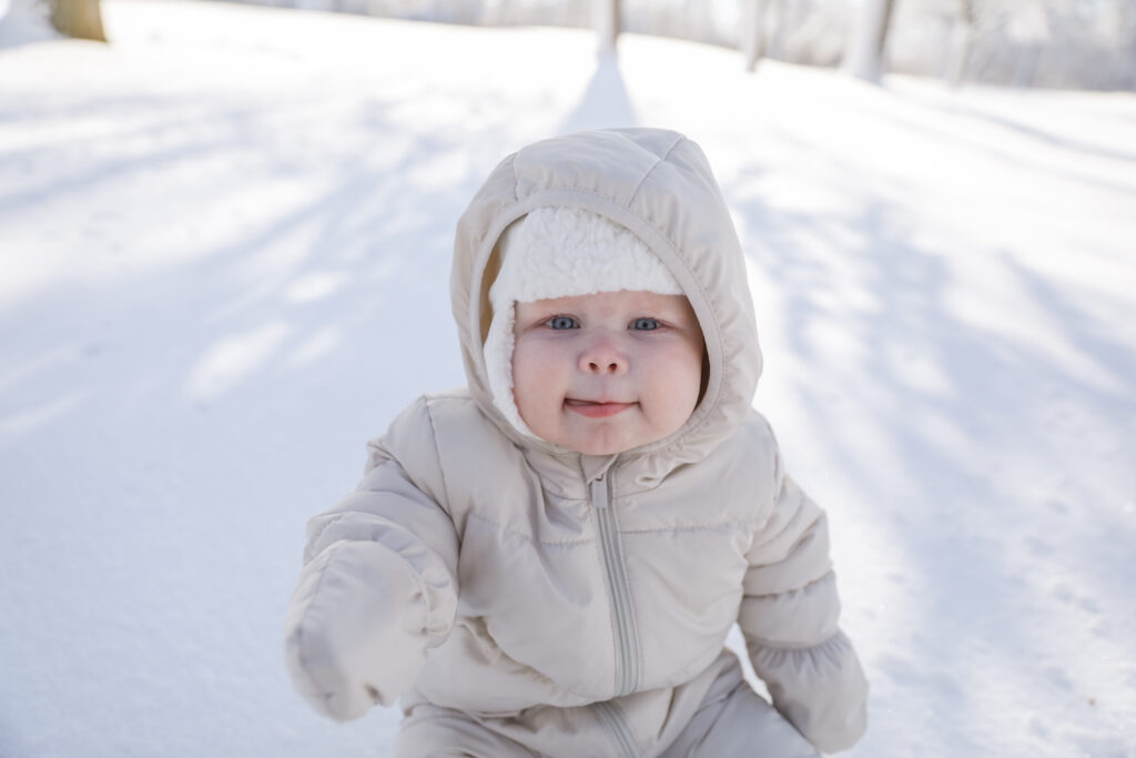 Baby girl, bundled in a white snowsuit, looks up to the camera and points and smiles in the snow on a bright day