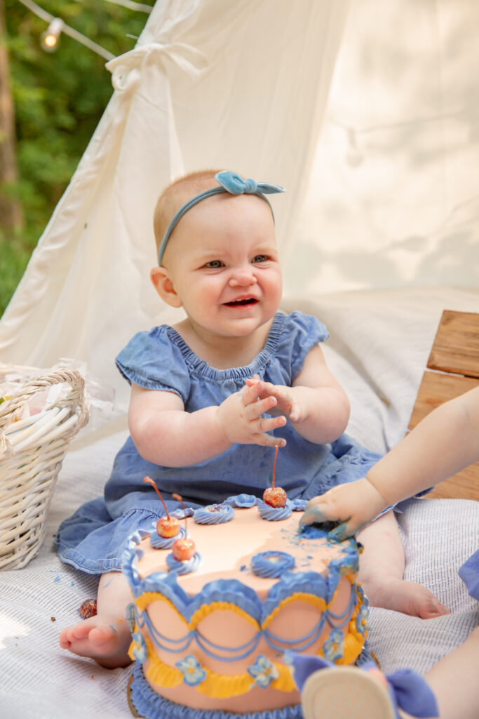 Baby girl in denim dress claps and smiles in front of a beautiful cake during cake smash photo shoot