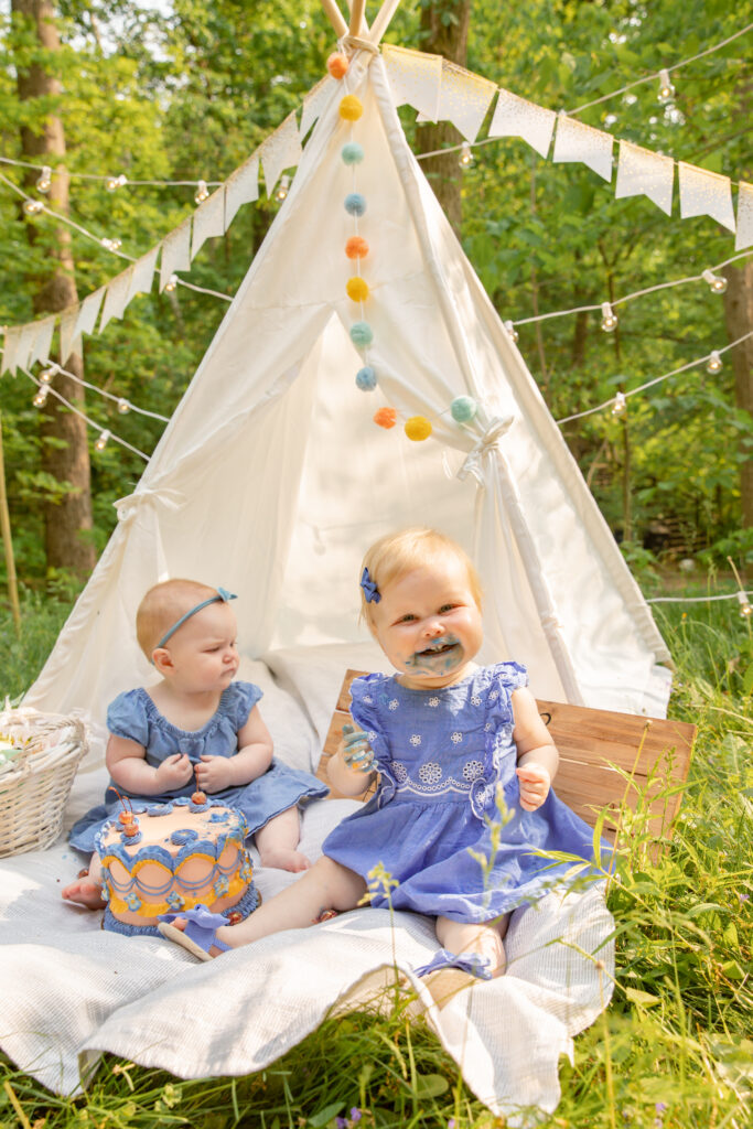 Two babies sit on a white blanket in front of a white teepee during a cake smash photo shoot with a beautiful classically decorated orange and purple cake made by Sweet Heather Anne