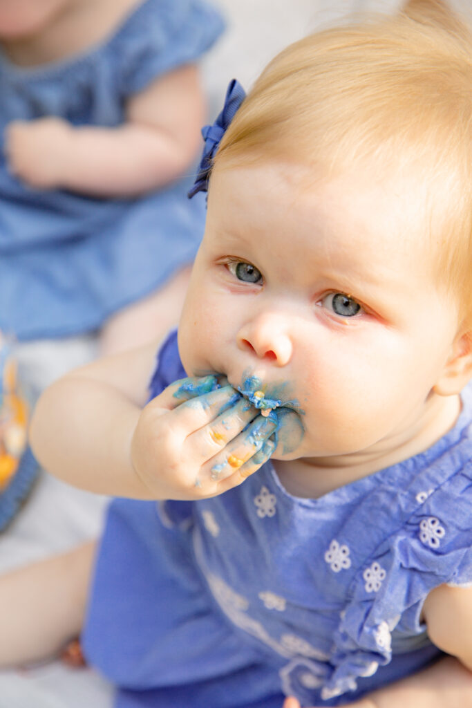 Close up of a baby girl with frosting on hands and mouth licking her fingers during a cake smash photoshoot