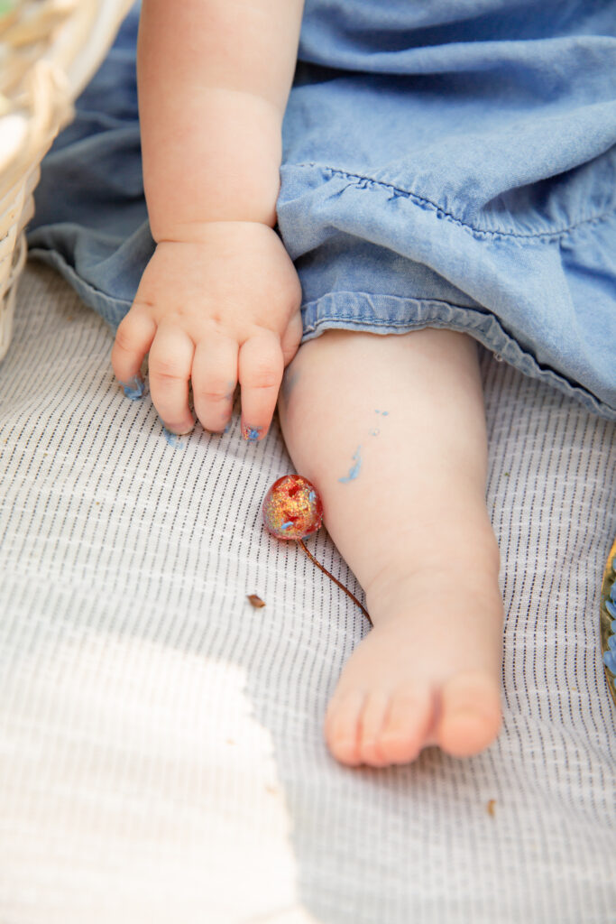 Close up of a baby's hand, leg and feet on a blanket during a cake smash session outdoors with a glittery cherry resting on her leg as she reaches for it