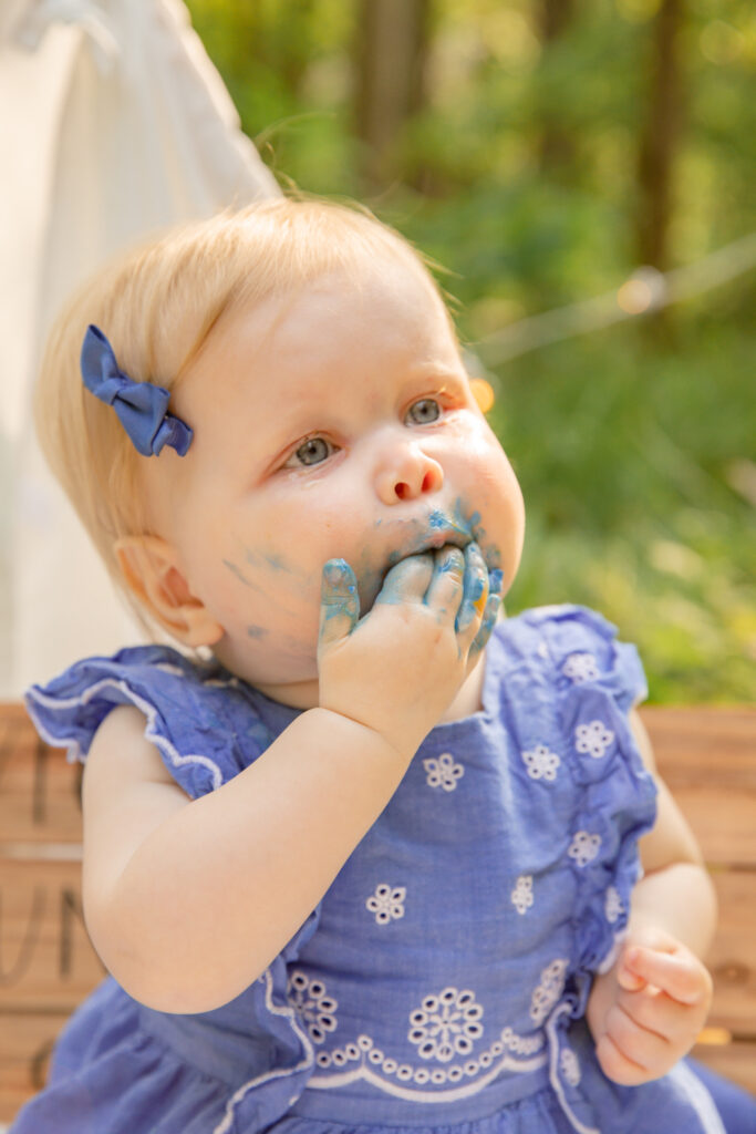 Close up of a baby girl with purple frosting on her hand and mouth, eating cake during an outdoor cake smash session