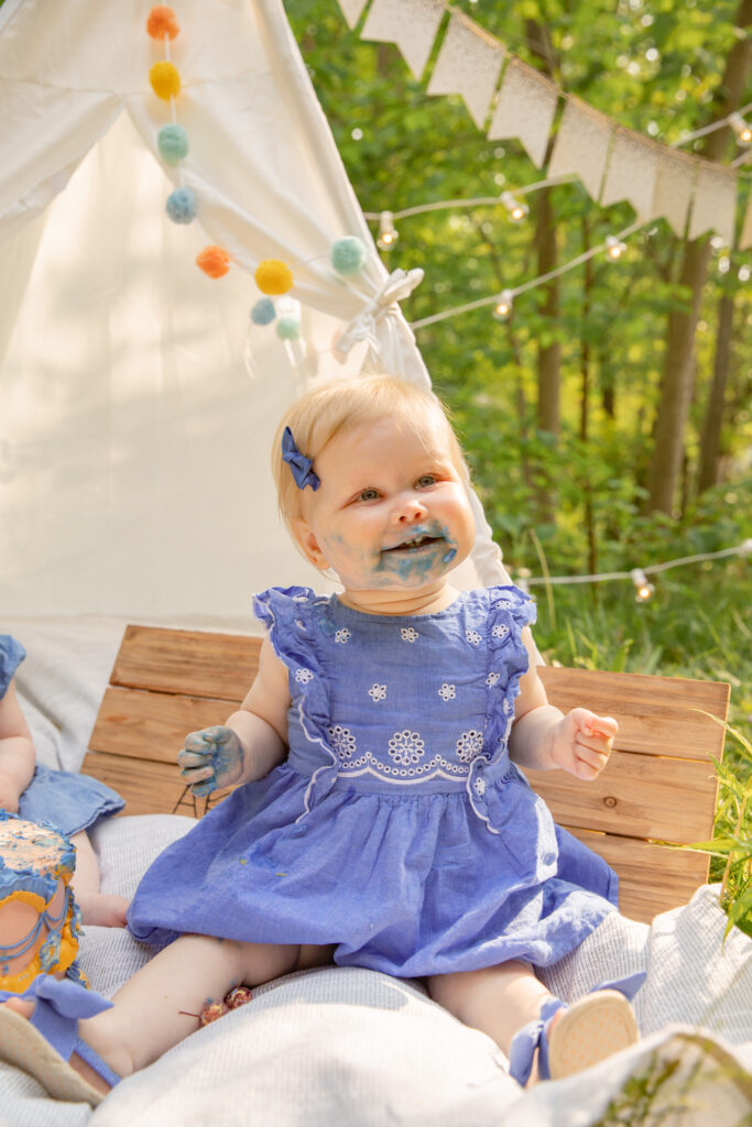 Baby girl in purple dress smiles off camera and sits on blanket in front of a teepee with purple frosting all over face and hands during cake smash photo shoot