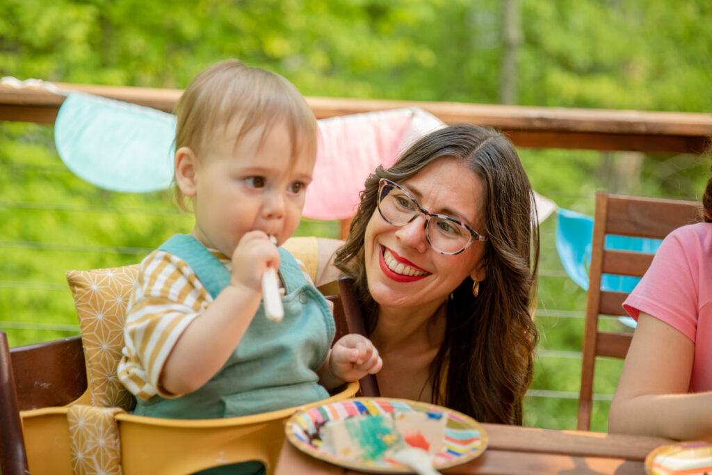 A mother smiles at her baby boy who intently is eating his chocolate birthday cake at an outdoor birthday party