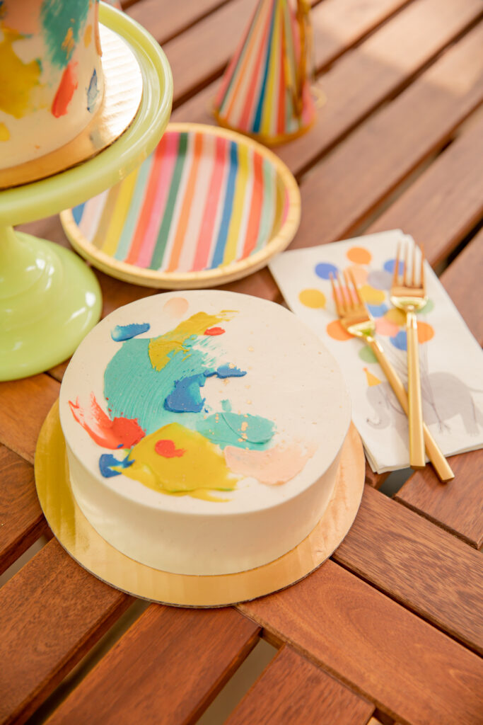 A beautiful cake by Sweet Heather Anne sits on a patio table with cute rainbow party supplies all around