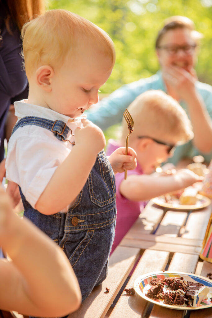 Toddler boy in overalls and white shirt stands at an outdoor table eating chocolate cake with other party guests in the background