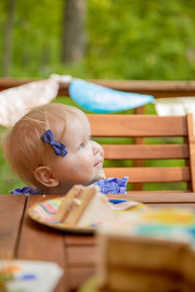 A baby girl looks off camera smiling with frosting on her face, purple bow in hair, at an outdoor party with birthday cake on a rainbow plate in front of her