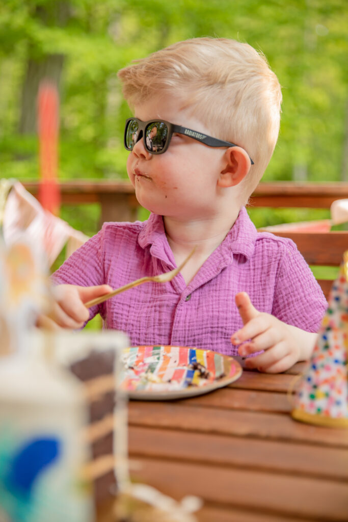 Young boy in bright purple shirt and black sunglasses finishes a piece of chocolate cake at an outdoor party