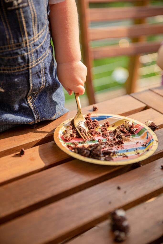 Close up of a toddler boy's hand with a fork eating the remains of a piece of chocolate cake off a rainbow paper plate outside