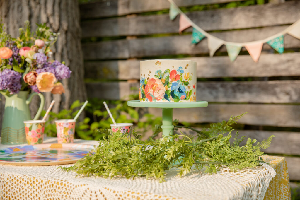 A beautiful cake by Sweet Heather Anne sits on a green milk glass stand with flowers and garden party decorations all around