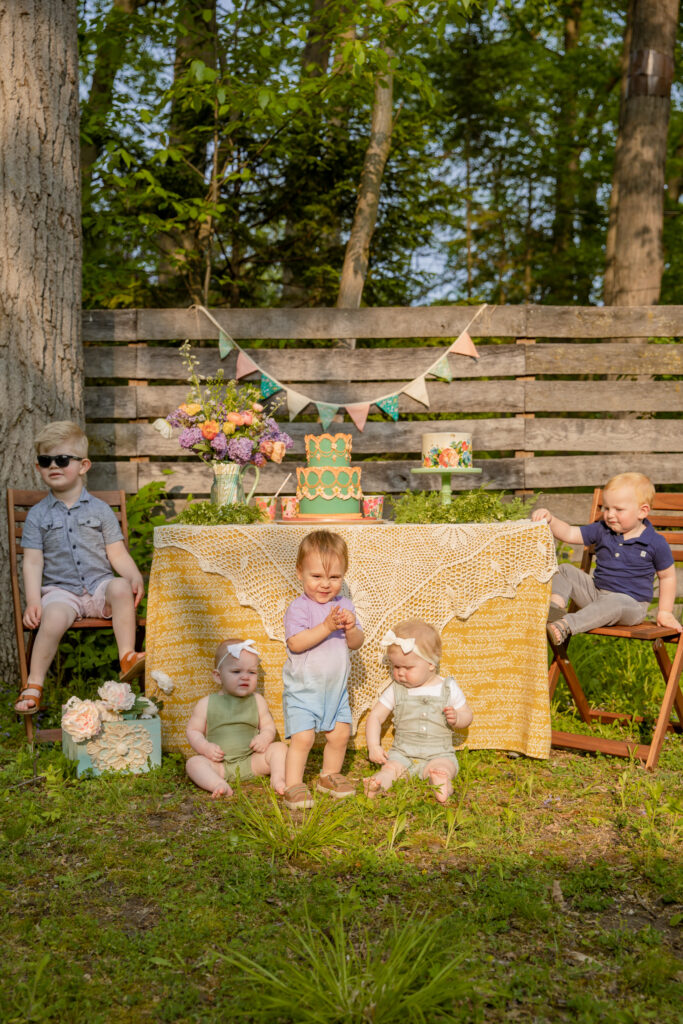 Toddlers and babies sit and stand in front of a table with a yellow cloth and lacy decoration draped over it and cakes and party decorations atop at an outdoor celebration