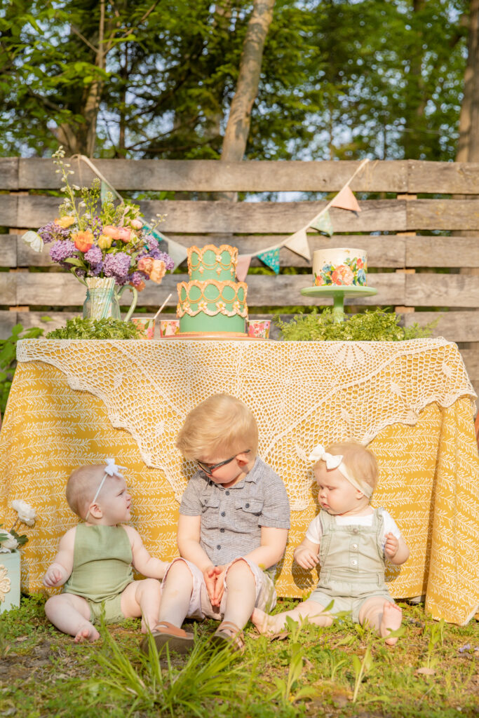 A toddler boy sits on the ground between two baby girls in front of a table with a yellow cloth and lacy decoration draped over it and cakes and party decorations atop at an outdoor celebration