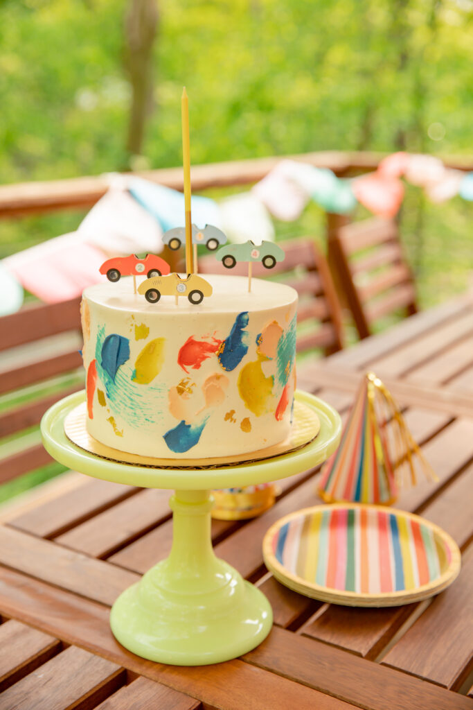 A beautiful cake by Sweet Heather Anne sits on a green milk glass stand with party hats and supplies sitting around