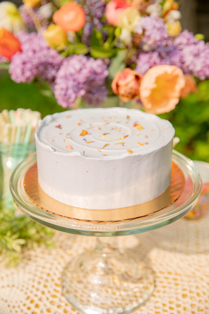 A beautiful cake by Sweet Heather Anne sits on a glass stand with flowers and paper straws on an outdoor table