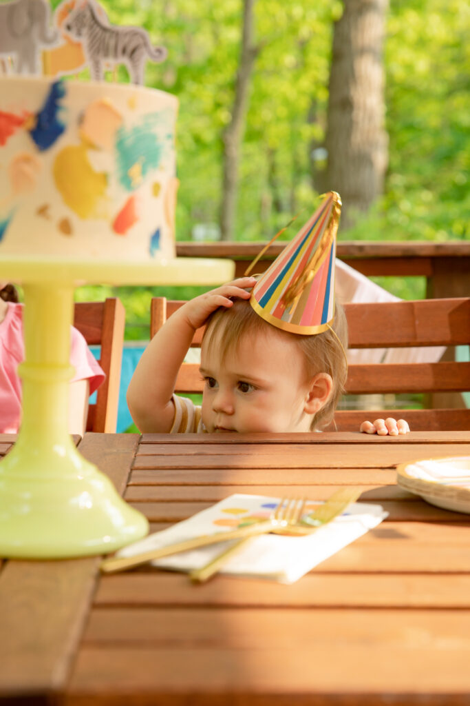 A young toddler sits at an outdoor table, face only just coming up over the top, wearing a party hat waiting for the birthday cake that sits in front of him on the table