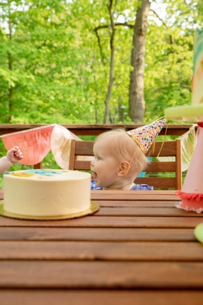 Baby girl wears a party hat and smiles big smile off camera with sweet little cake on outdoor table at birthday party