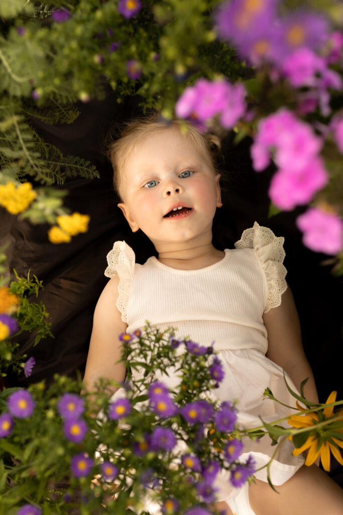 toddler girl lays amongst yellow and purple flowers and looks up towards camera