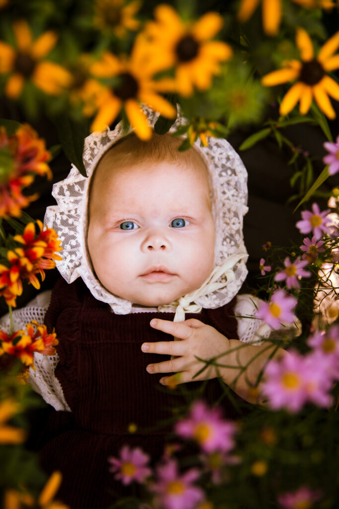 baby girl with lace bonnet lays amongst yellow, pink, and orange flowers while looking up at camera