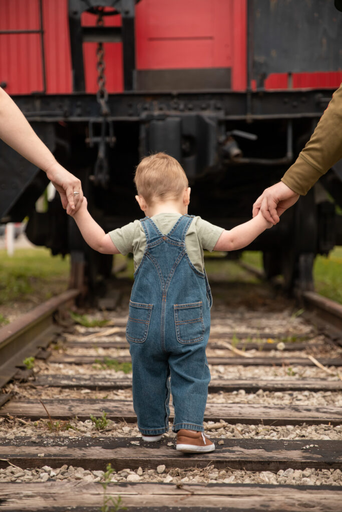 toddler boy walks away from camera towards caboose at outdoor train museum, wearing overalls, holding parents hands