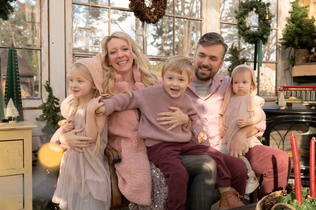 A family with three children sit together smiling and laughing at the camera in a greenhouse decorated for Christmas