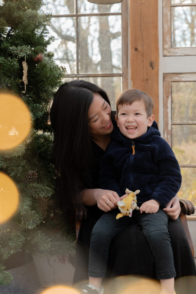 A young boy sits on the lap of his mother and laughs in a greenhouse decorated for christmas