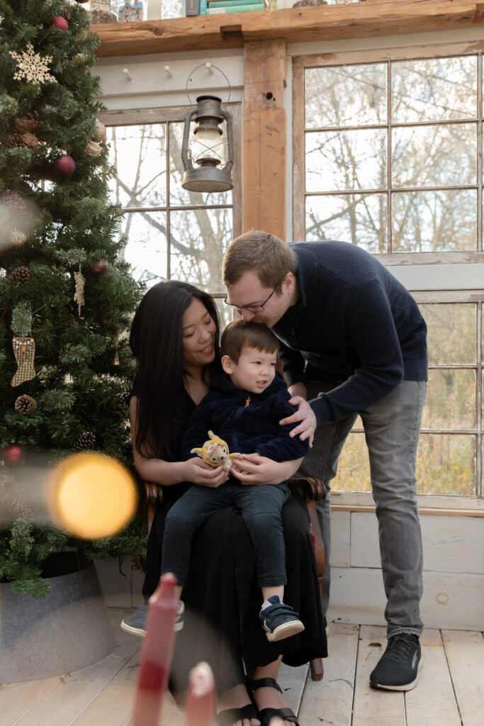 A mother, father, and young son sit on a chair and laugh together in a greenhouse decorated for christmas