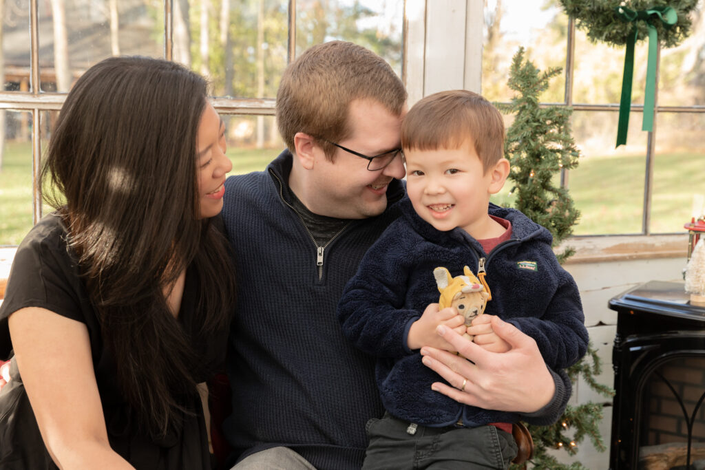 A mother and father sit with their young son, the father snuggles into the boy's head while the boy smiles at camera and the mother smiles at the boy in a greenhouse decorated for Christmas.