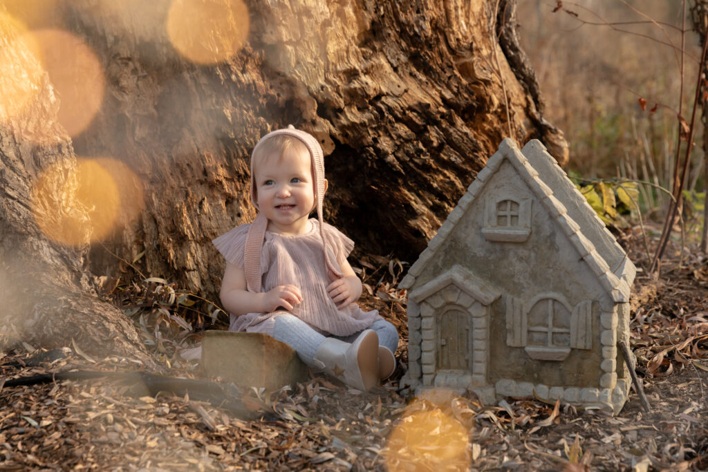 A young toddler girl sits between a large tree and a small concrete playhouse, smiling off camera with twinkle lights around