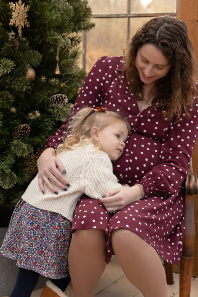 Young girl snuggles with mama's pregnant belly in a greenhouse decorated for christmas