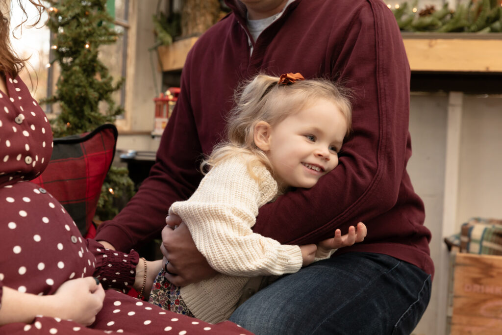 Young girl snuggles in dad's arms and smiles in a greenhouse decorated for Christmas