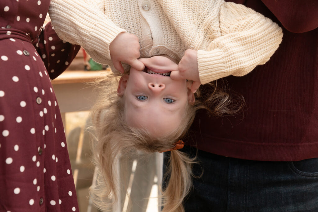 Young toddler girl hangs upside down and makes a silly face in a greenhouse decorated for Christmas