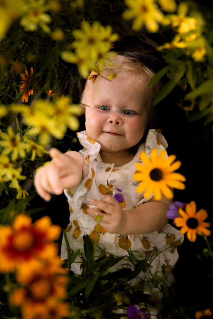 baby girl lays amongst yellow and orange flowers and points up towards camera