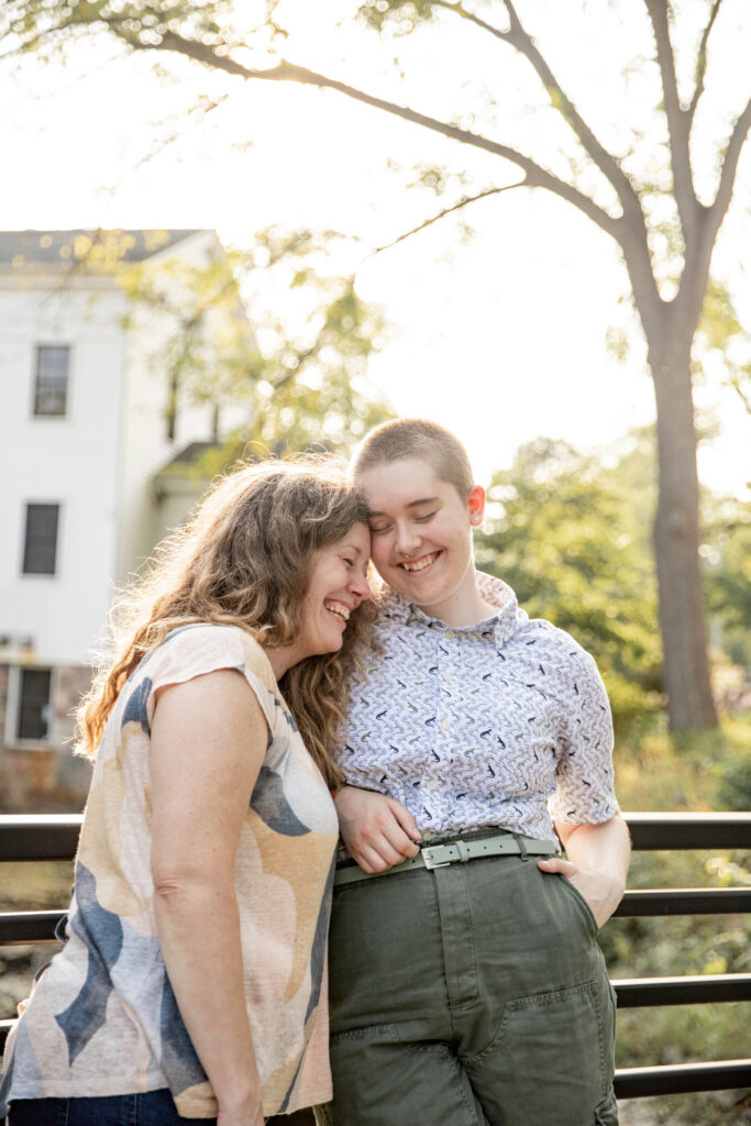 mother and adult child laugh and snuggle together on bridge during golden hour at Sharon Mills Park in Manchester Michigan