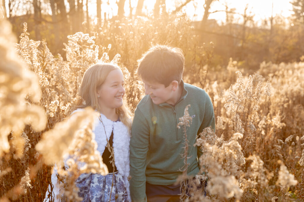 Siblings stand in a field of tall grass during golden hour and laugh together