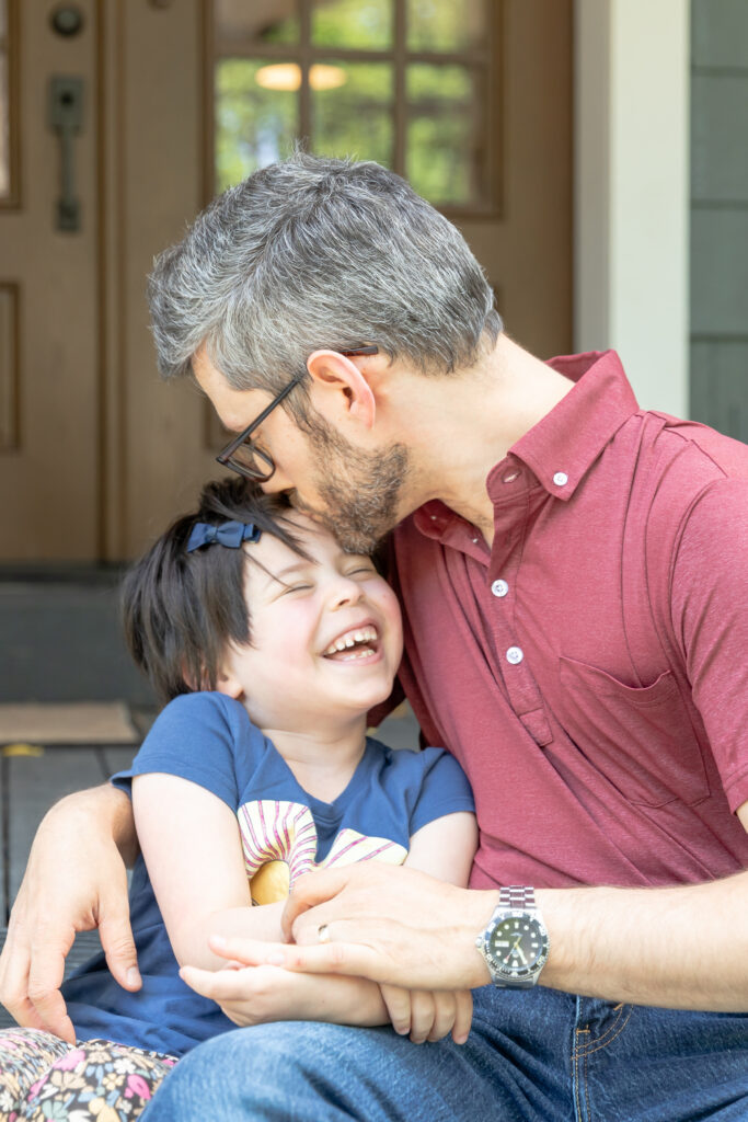 Father and young daughter sit on porch of family home while dad leans down to kiss girl on her forehead while she laughs during in-home session in ann arbor