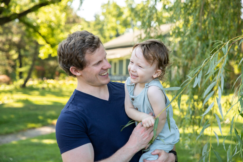 father holds toddler boy under willow tree and tickles him while they both laugh at white lotus farms in ann arbor