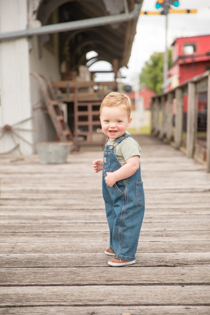 toddler boy stands on boardwalk at train depot museum in saline Michigan and looks at camera with huge smile