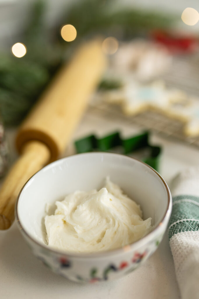 cookie frosting in small christmas bowl with rolling pin, cookie cutter, cookies, and greenery with twinkle lights in background