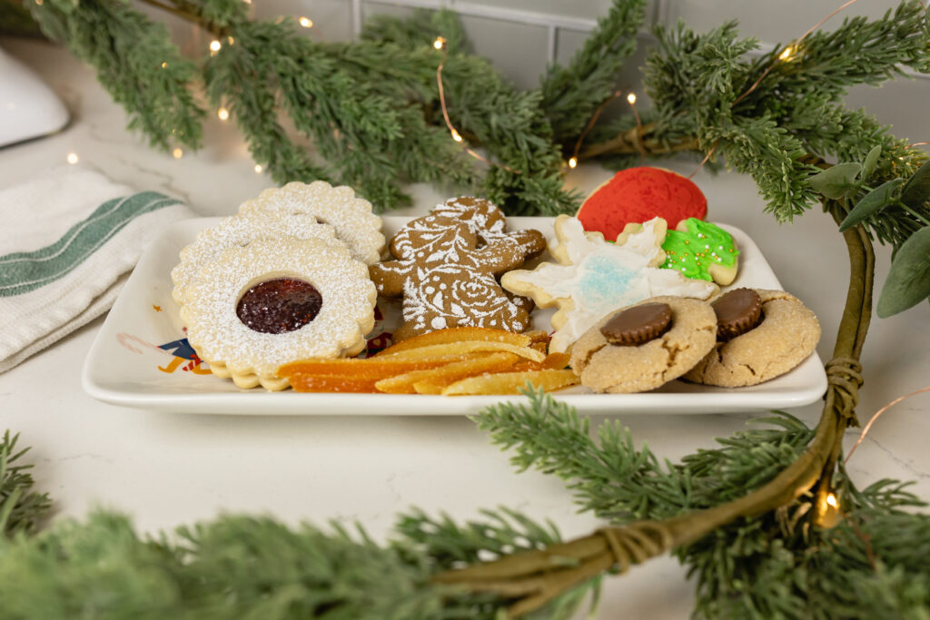 linzer cookies, gingerbread cookies, candied orange peel, cut out cookies, and peanut butter blossoms sitting on platter with christmas greenery and twinkle lights