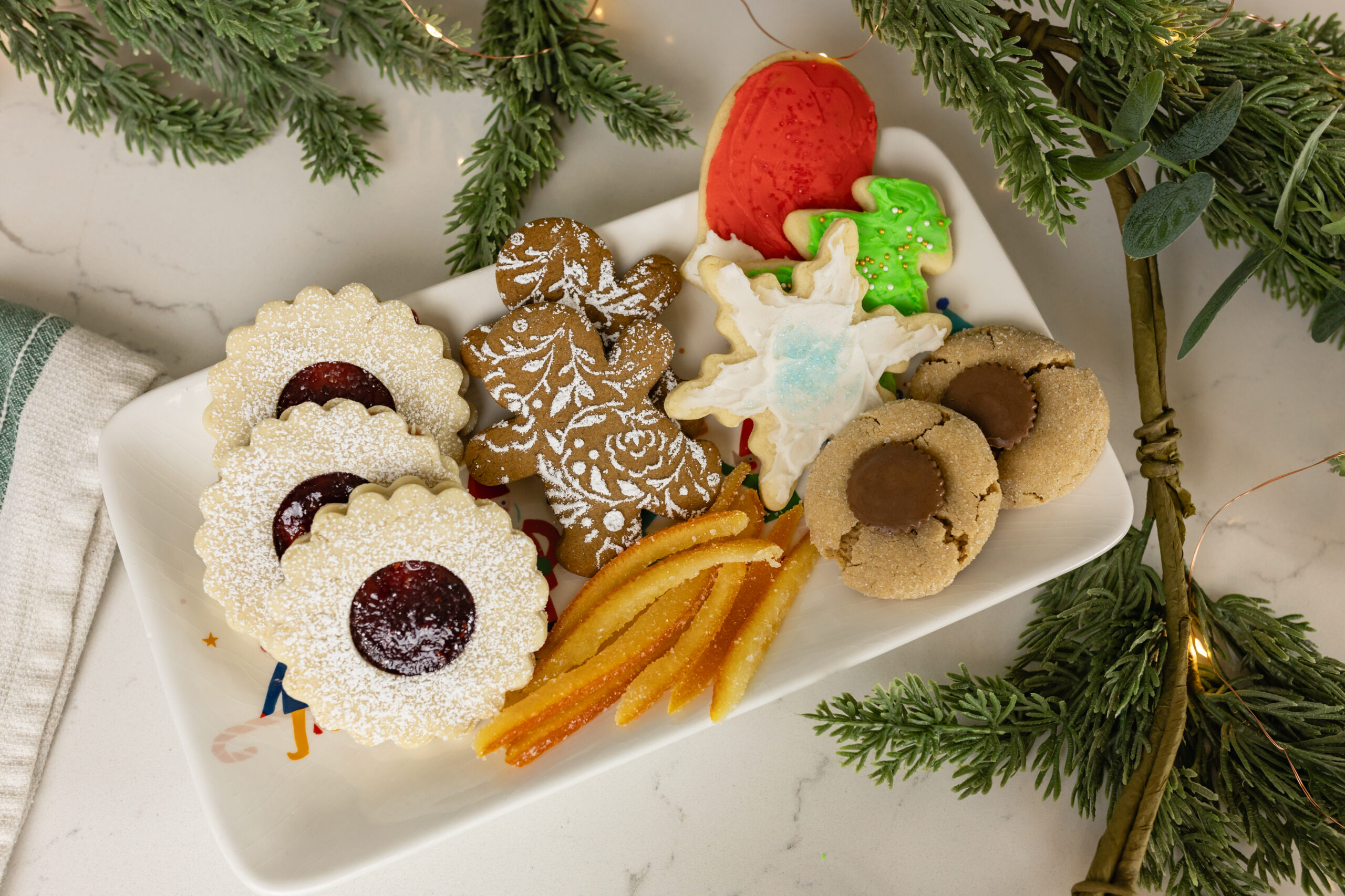 linzer cookies, gingerbread cookies, candied orange peel, cut out cookies, and peanut butter blossoms sitting on platter with christmas greenery and twinkle lights