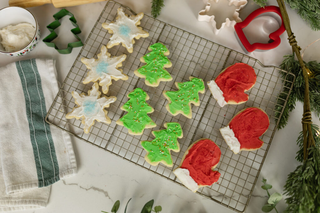 christmas cut out sugar cookies decorated on rack with rolling pin, cookie cutters, frosting, twinkle lights, and christmas greenery around