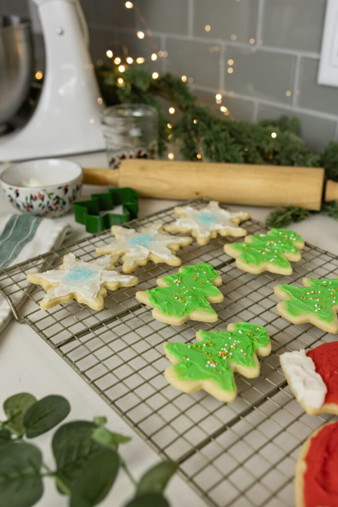 christmas cut out sugar cookies decorated on rack with rolling pin, cookie cutters, frosting, twinkle lights, and christmas greenery around