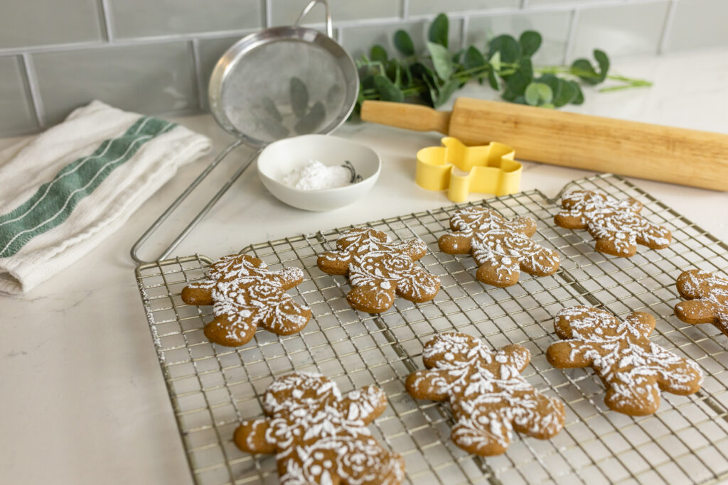 gingerbread cookies decorated with lacy powdered sugar on top sit on rack with rolling pin, small bowl, sifter, cookie cutter, and greenery around