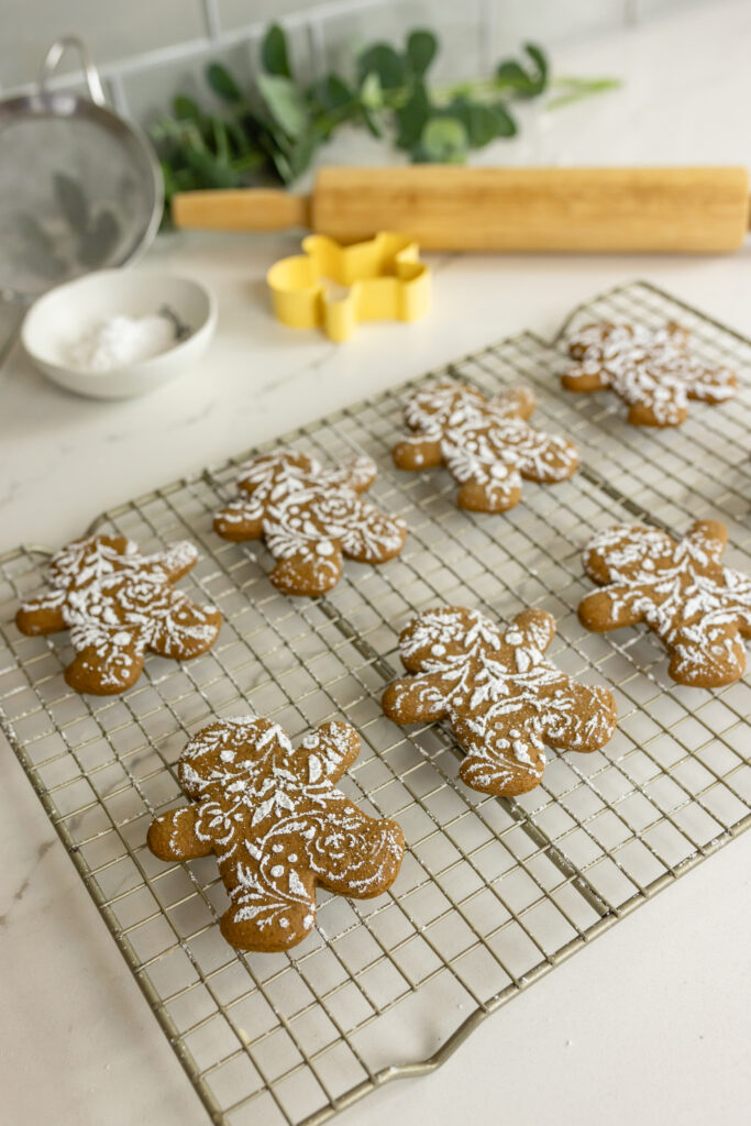 gingerbread cookies decorated with lacy powdered sugar on top sit on rack with rolling pin, small bowl, sifter, cookie cutter, and greenery around