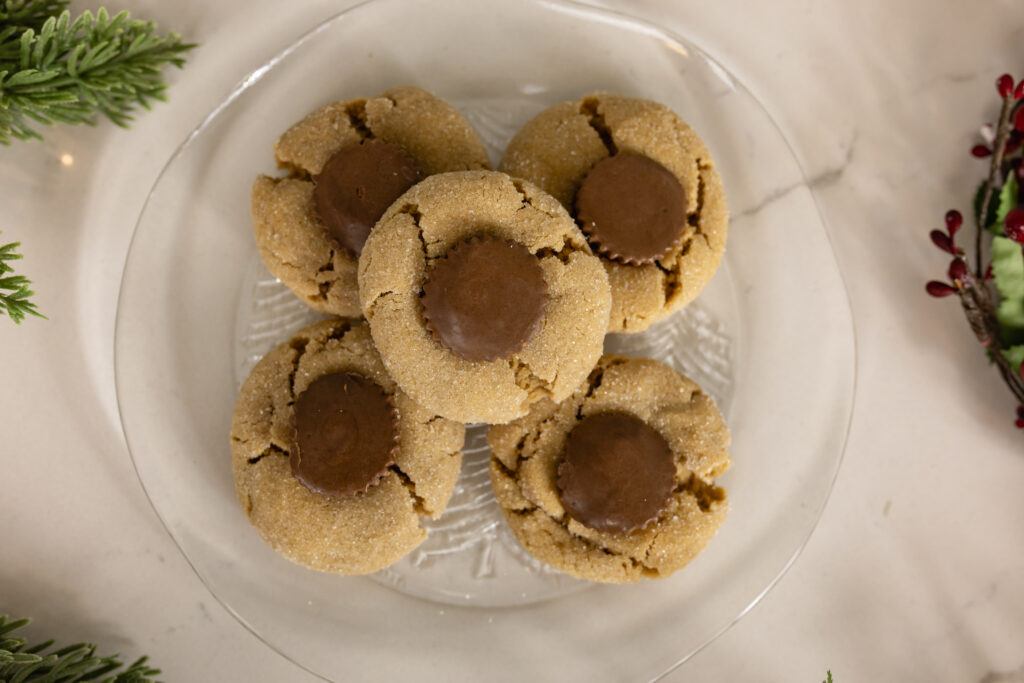 peanut butter blossoms sitting on clear glass plate surrounded by christmas greenery