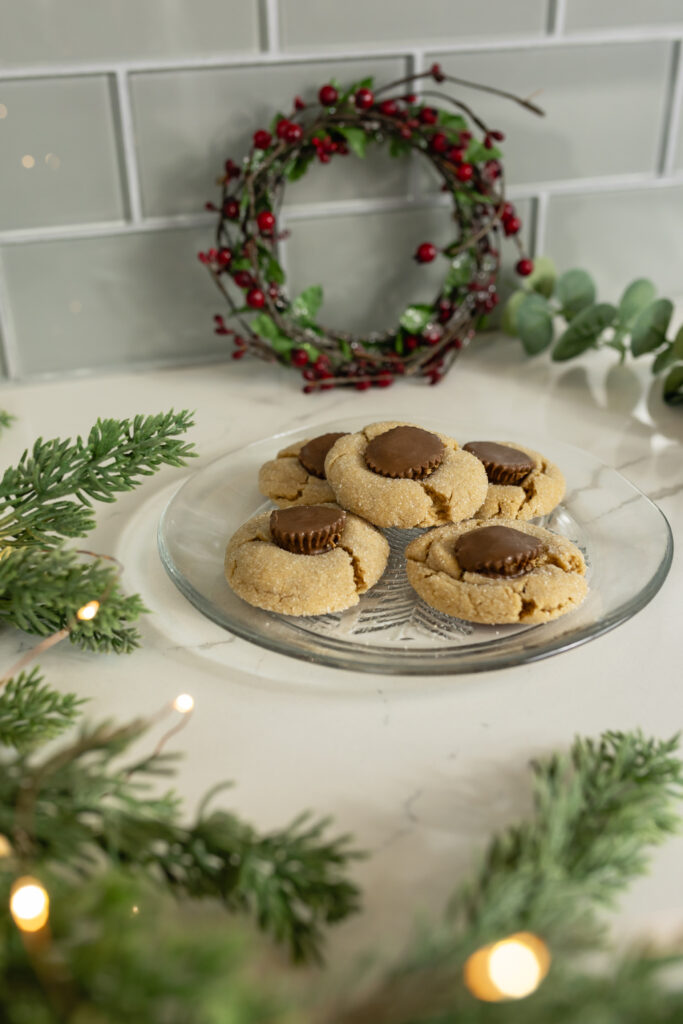 peanut butter blossoms sitting on clear glass plate surrounded by christmas greenery